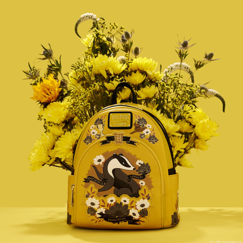 Yellow Hufflepuff mini backpack featuring the Hufflepuff badger on the front pocket, surrounded by white and grey flowers, sitting against a yellow background in front of a real bouquet of yellow flowers